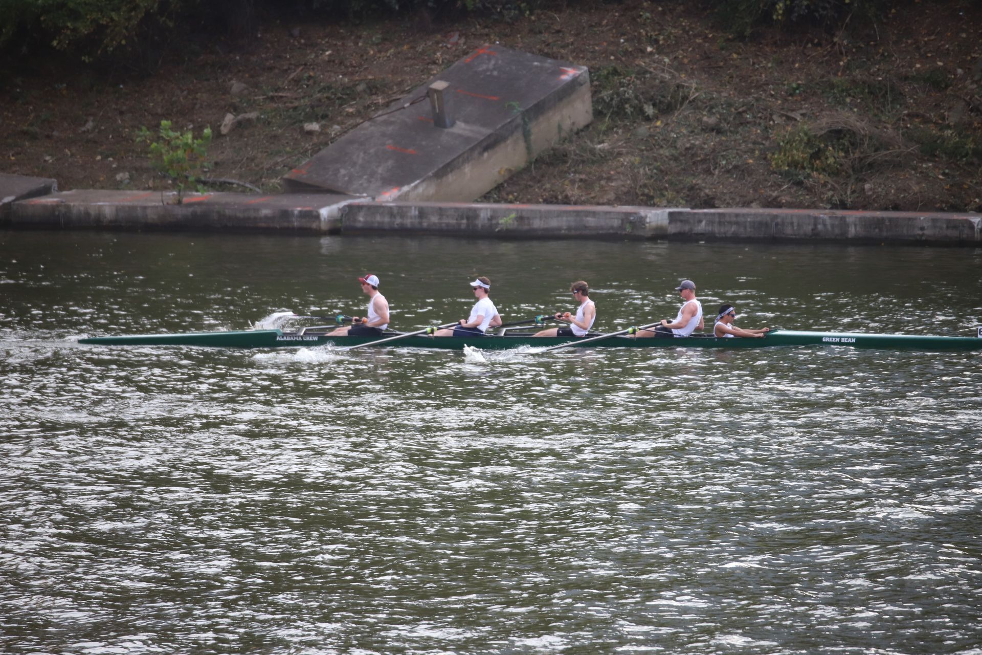 A rowing crew of four, wearing white, rows on a river. A concrete wall and building are in the background.