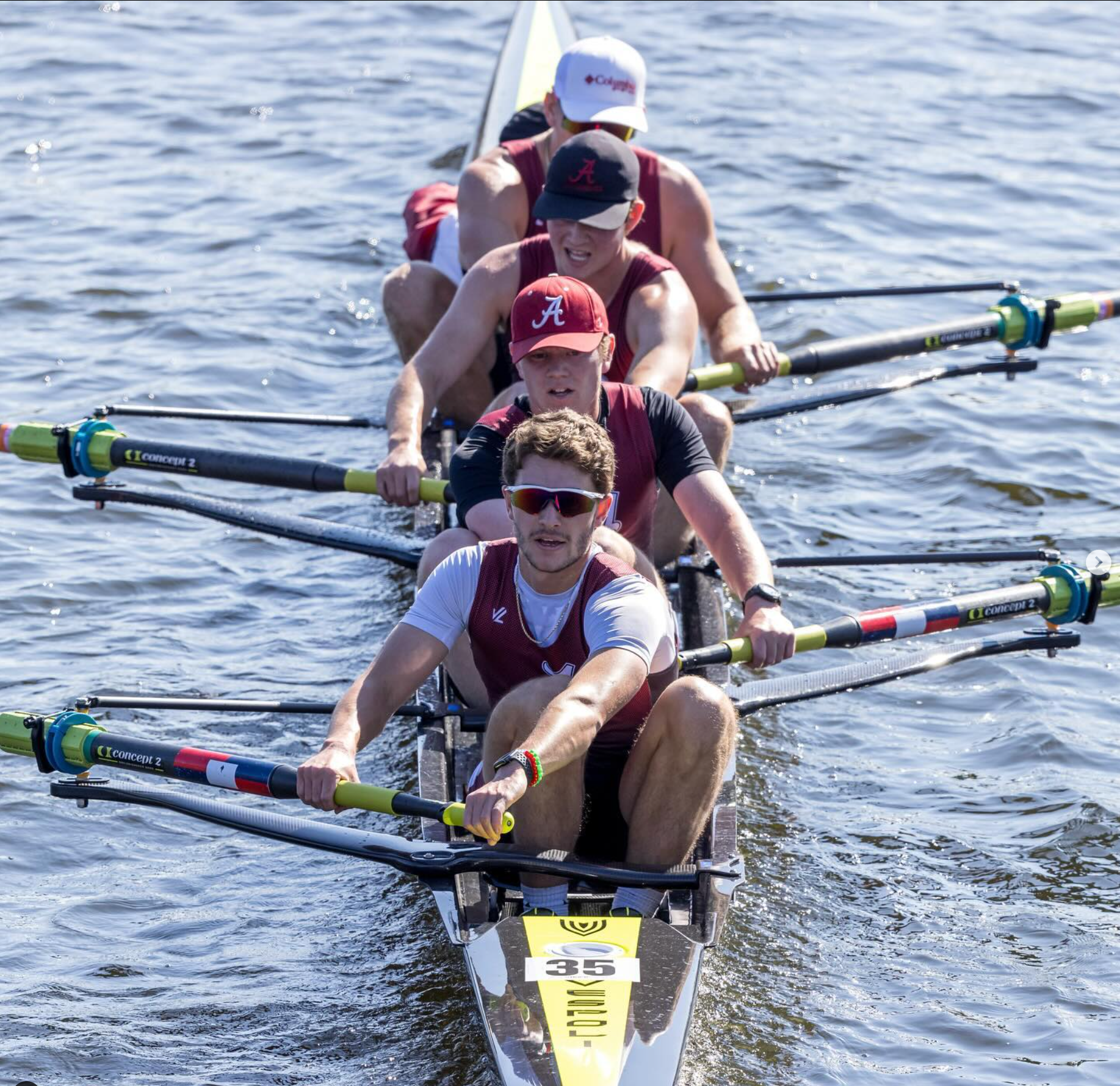 A rowing crew wearing burgundy uniforms. The rowers are actively pulling their oars, with the sun reflecting off the water.