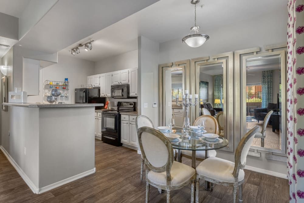 Modern open-plan kitchen with white cabinets and stainless steel appliances, adjacent to a dining area with a glass-top table and decorative chandelier, leading to a living room visible through mirrored sliding doors at Alante at the Islands in Chandler, AZ.