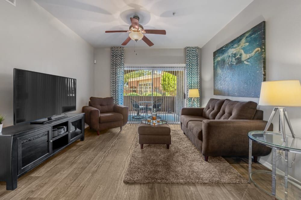 Modern living room interior with a large wall-mounted television, a brown sofa and armchair set, a glass-top side table with a lamp, and a cream-colored area rug. A large abstract painting adorns the wall, and patterned curtains frame a balcony door with a view of trees outside at Alante at the Islands in Chandler, AZ.