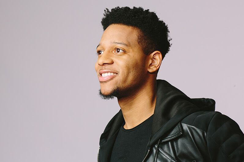 A young man wearing a black jacket and a black shirt is smiling.