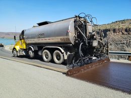 Asphalt distributor truck spraying sealant on a highway, roadside view with water and hills in the background.