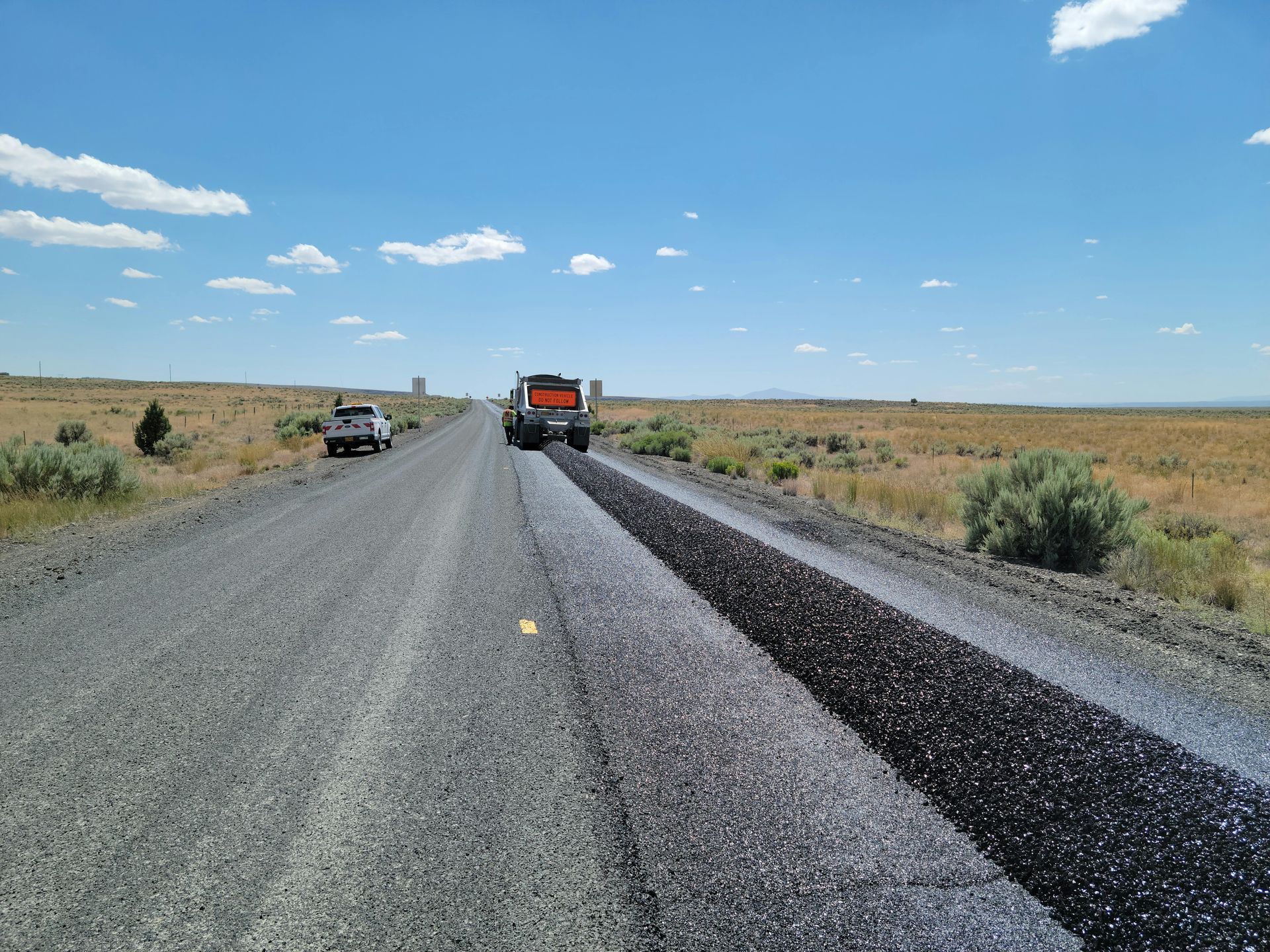 Road resurfacing in progress on a rural road under a blue sky; an orange truck and white pickup.