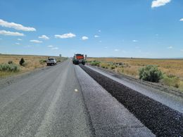 Road paving in progress, a truck applying asphalt on a rural road under blue sky.