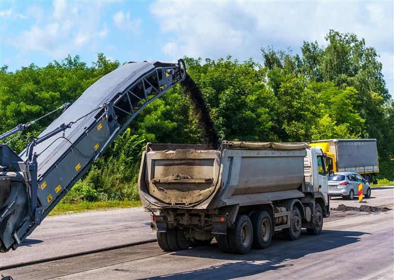 Road milling machine loading asphalt into a dump truck on a sunny road with trees.