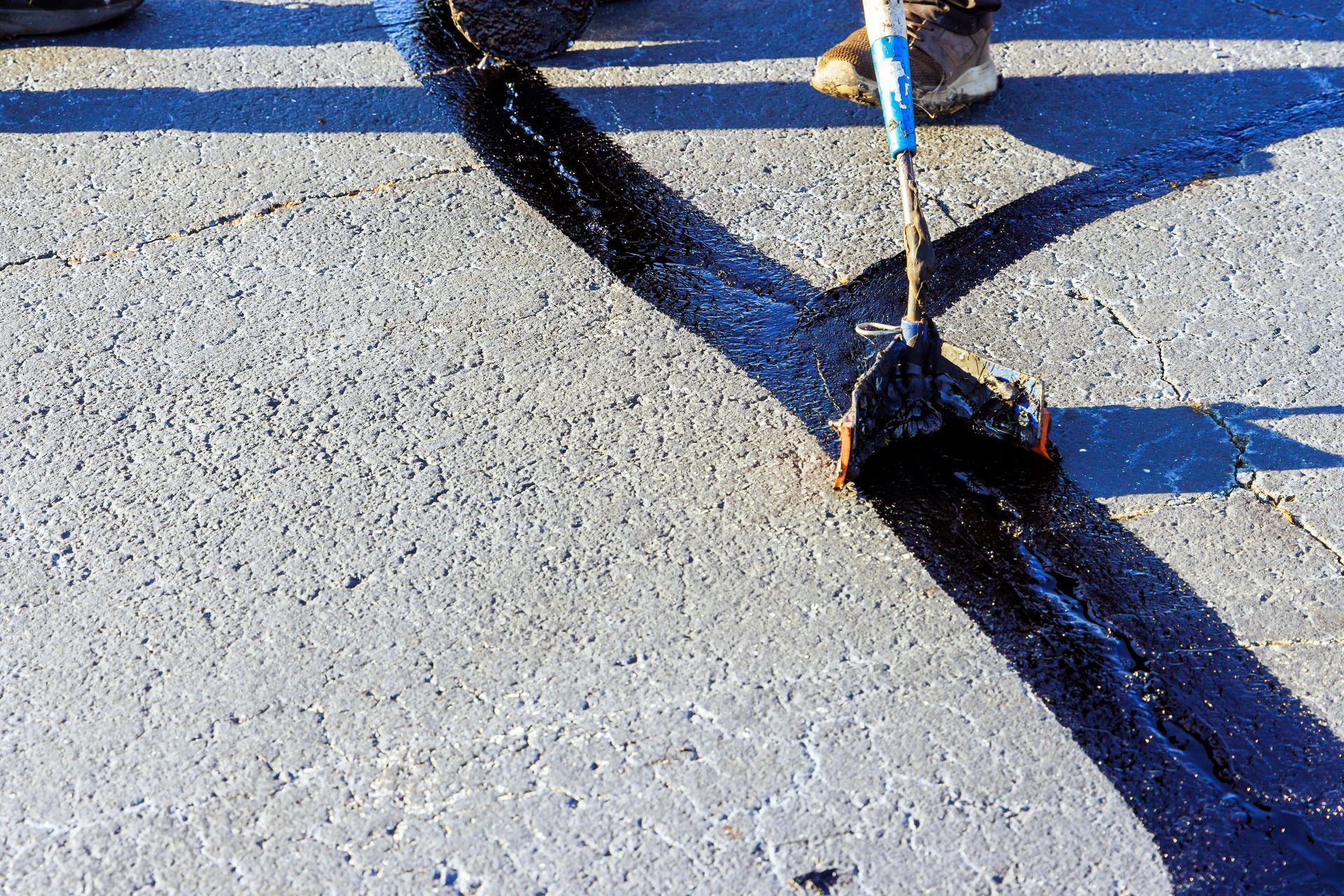 A person applying black sealant to a cracked asphalt surface with a roller.