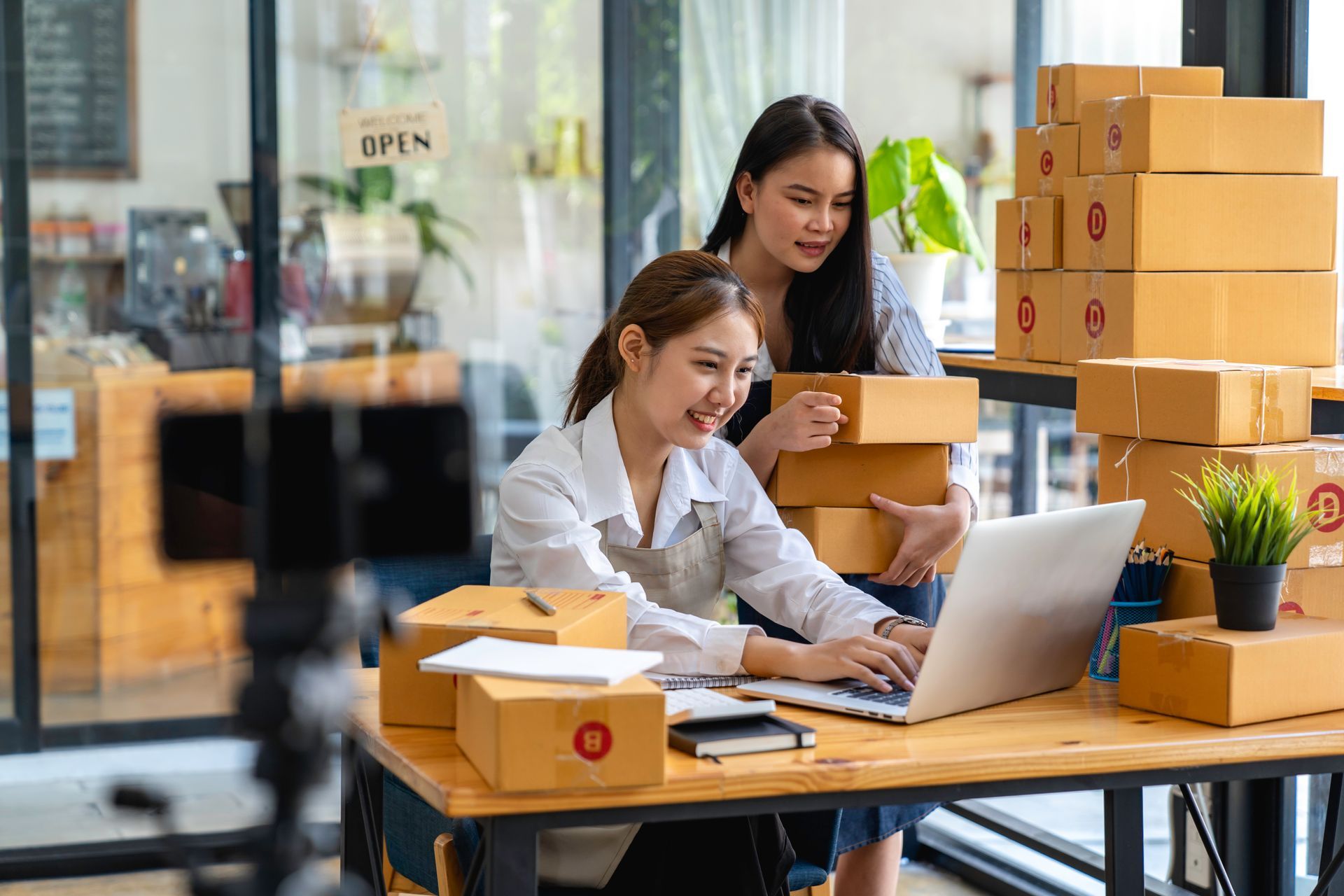 Two women are sitting at a desk with boxes and a laptop.