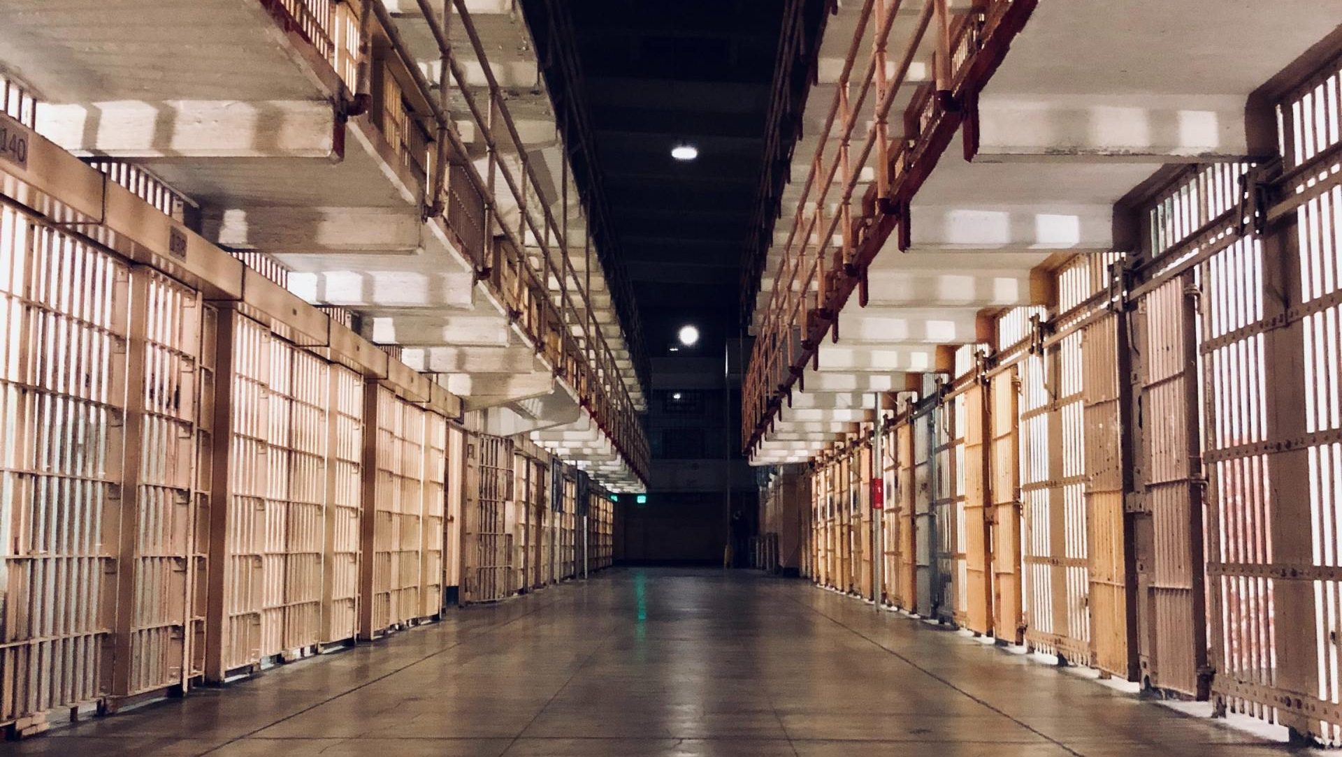 Interior view of a prison corridor with barred cells on either side. Dim lighting and a dark ceiling.