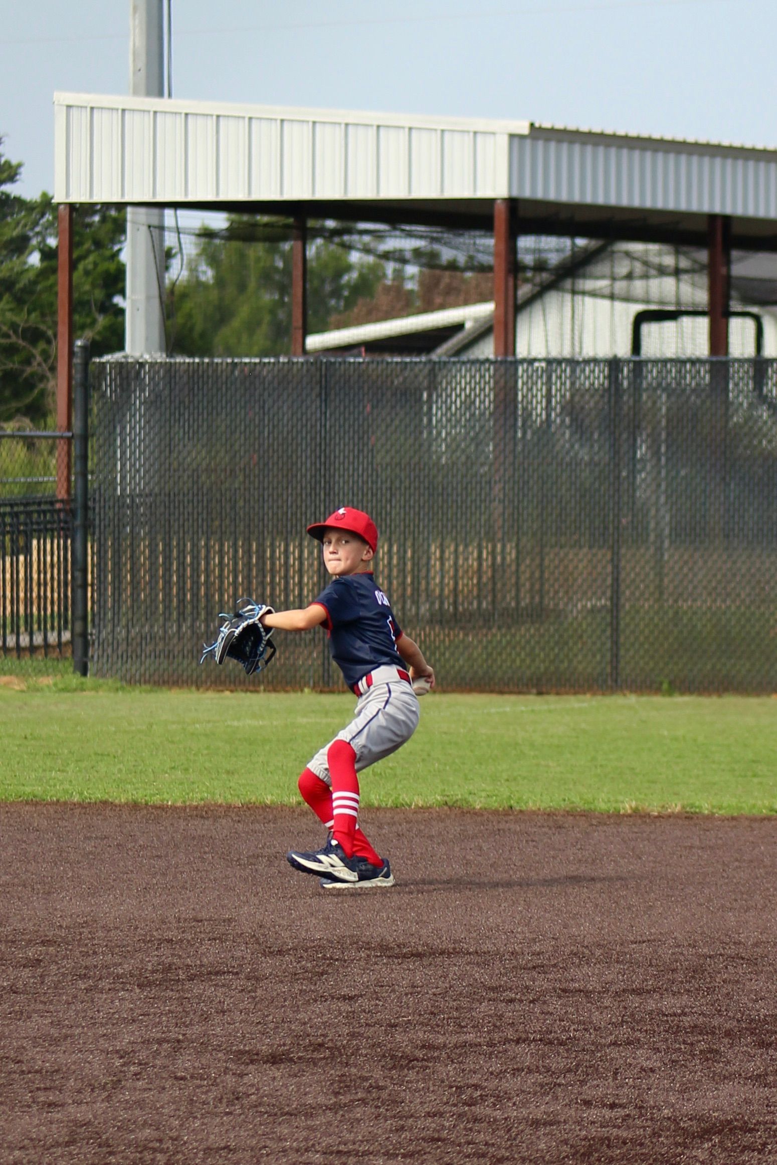 Young baseball pitcher in blue uniform winding up to throw a pitch on a dirt mound.