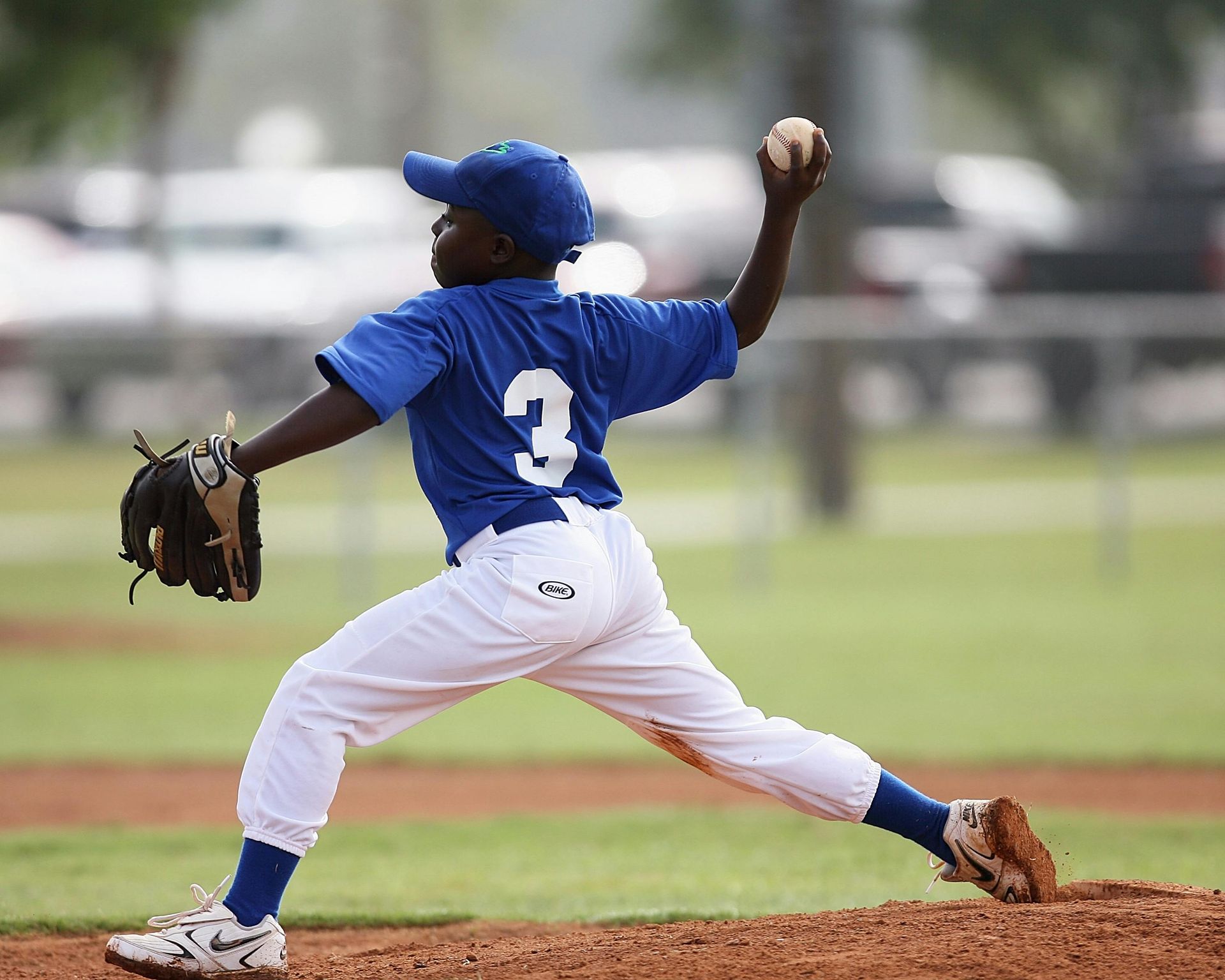 Young baseball pitcher in blue uniform winding up to throw a pitch on a dirt mound.