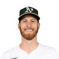 A baseball player wearing an Oakland Athletics cap smiles, with a full brown beard.