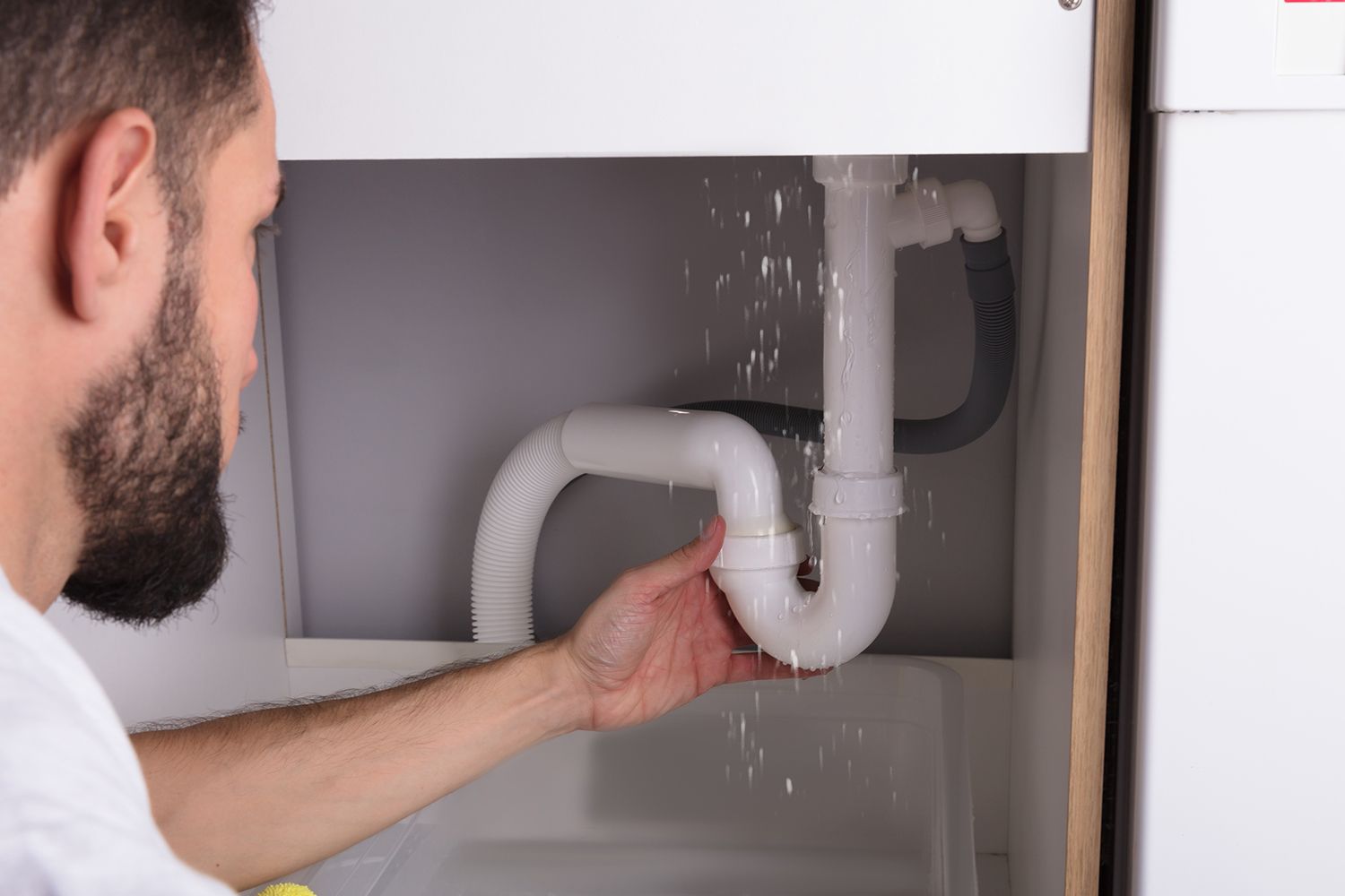A plumbing contractor fixing a leaking sink drainpipe under a cabinet.