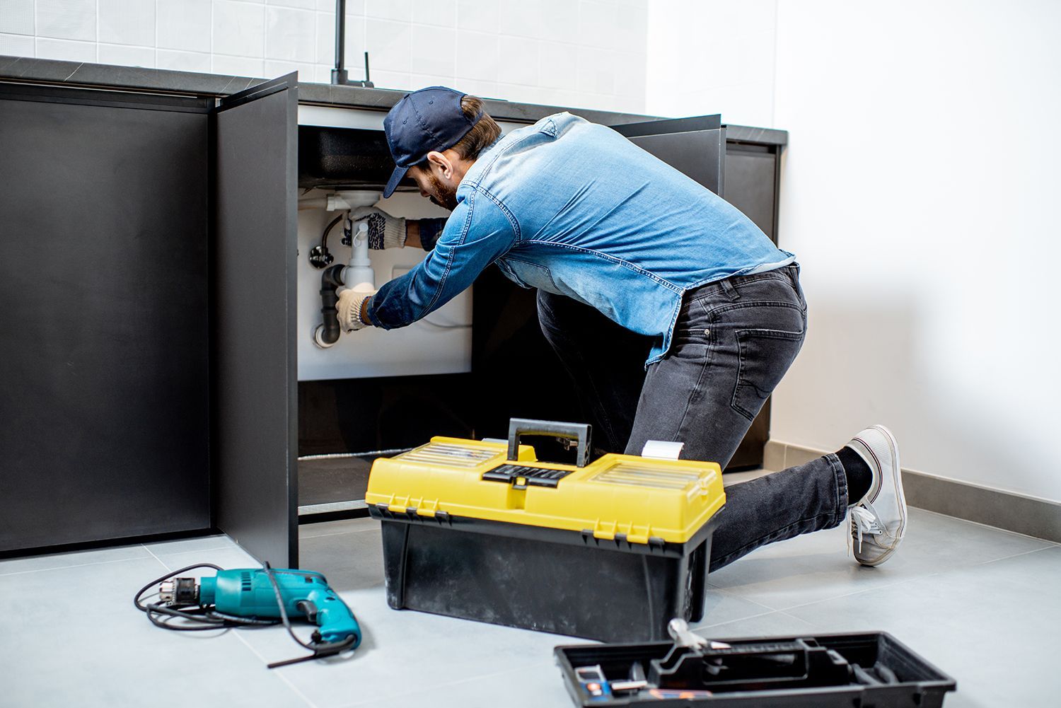 Plumber in blue shirt and cap mounting or repairing kitchen furniture in an apartment.