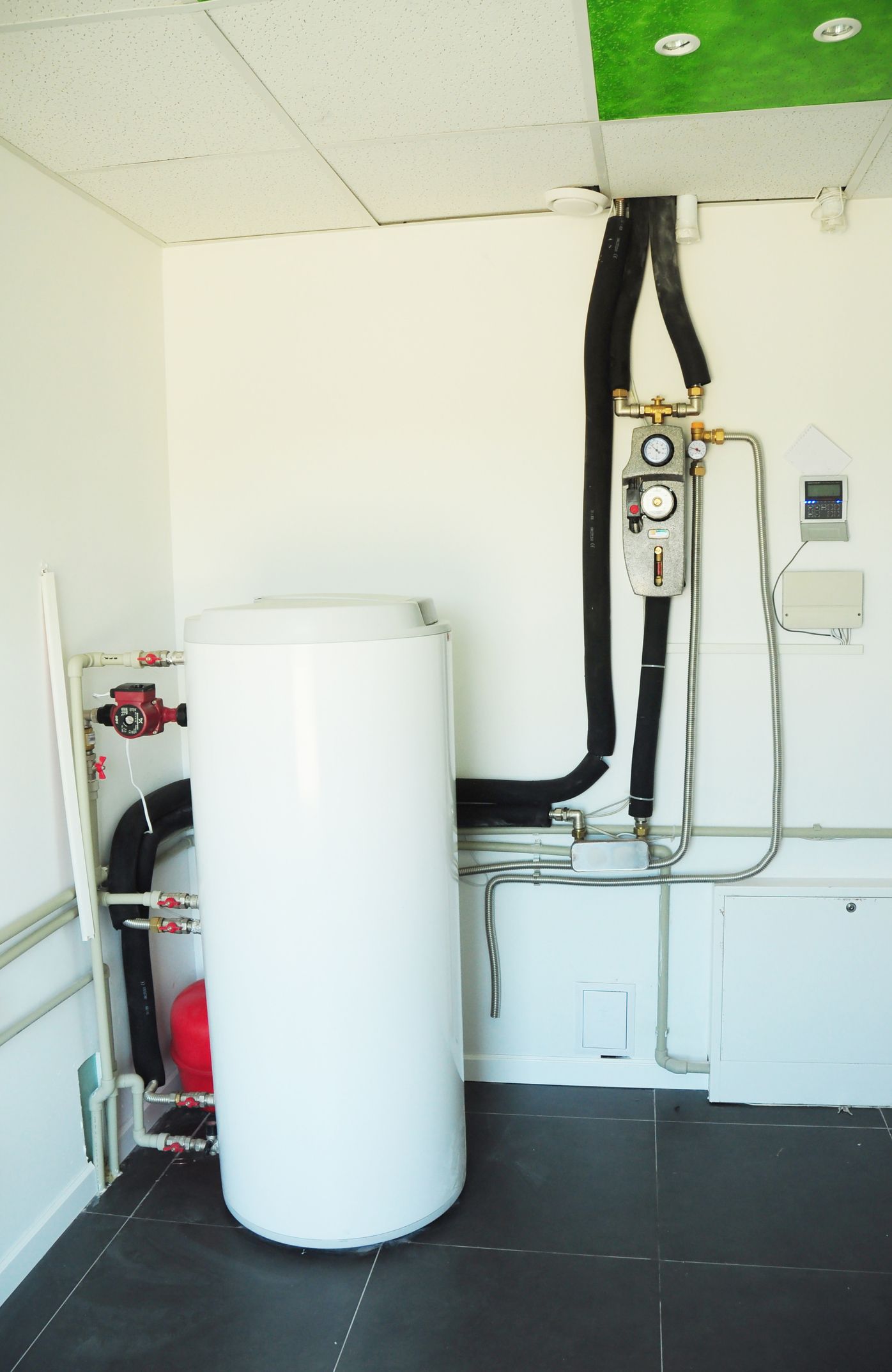 White water heater in a utility room, with black pipes and a control panel on the wall