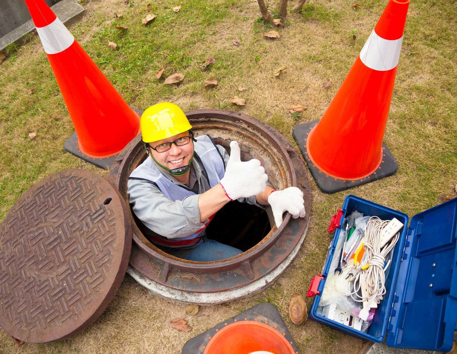 A worker in a manhole gives a thumbs up, with safety cones, a tool kit, and an open manhole cover