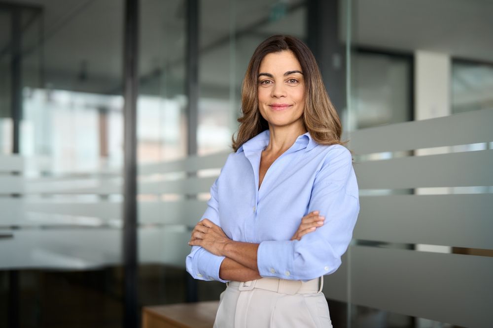 A woman in a blue shirt is standing in an office with her arms crossed.