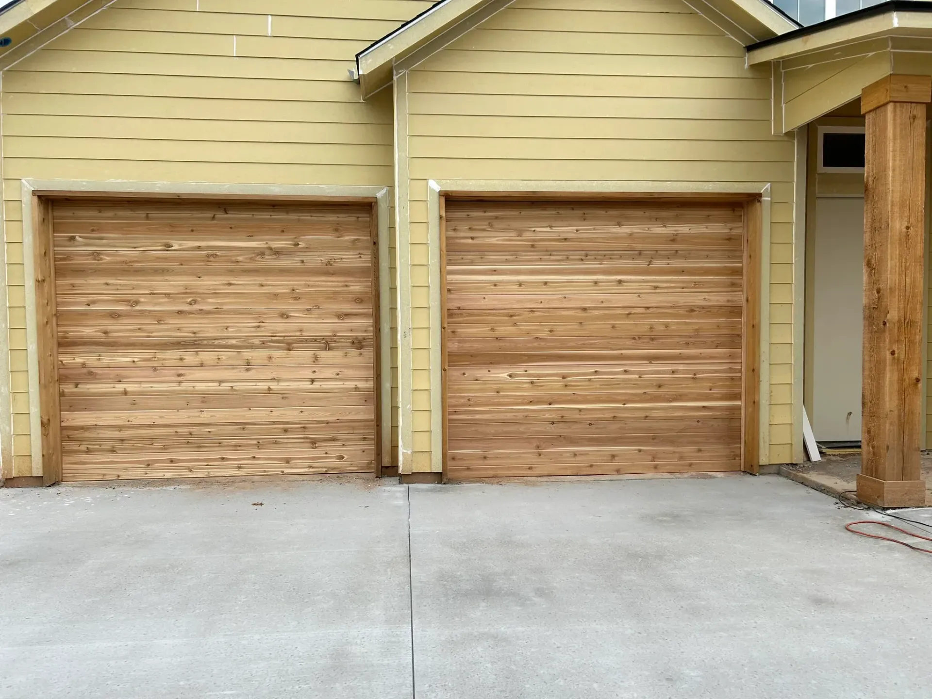 Two wooden garage doors are in front of a yellow house.