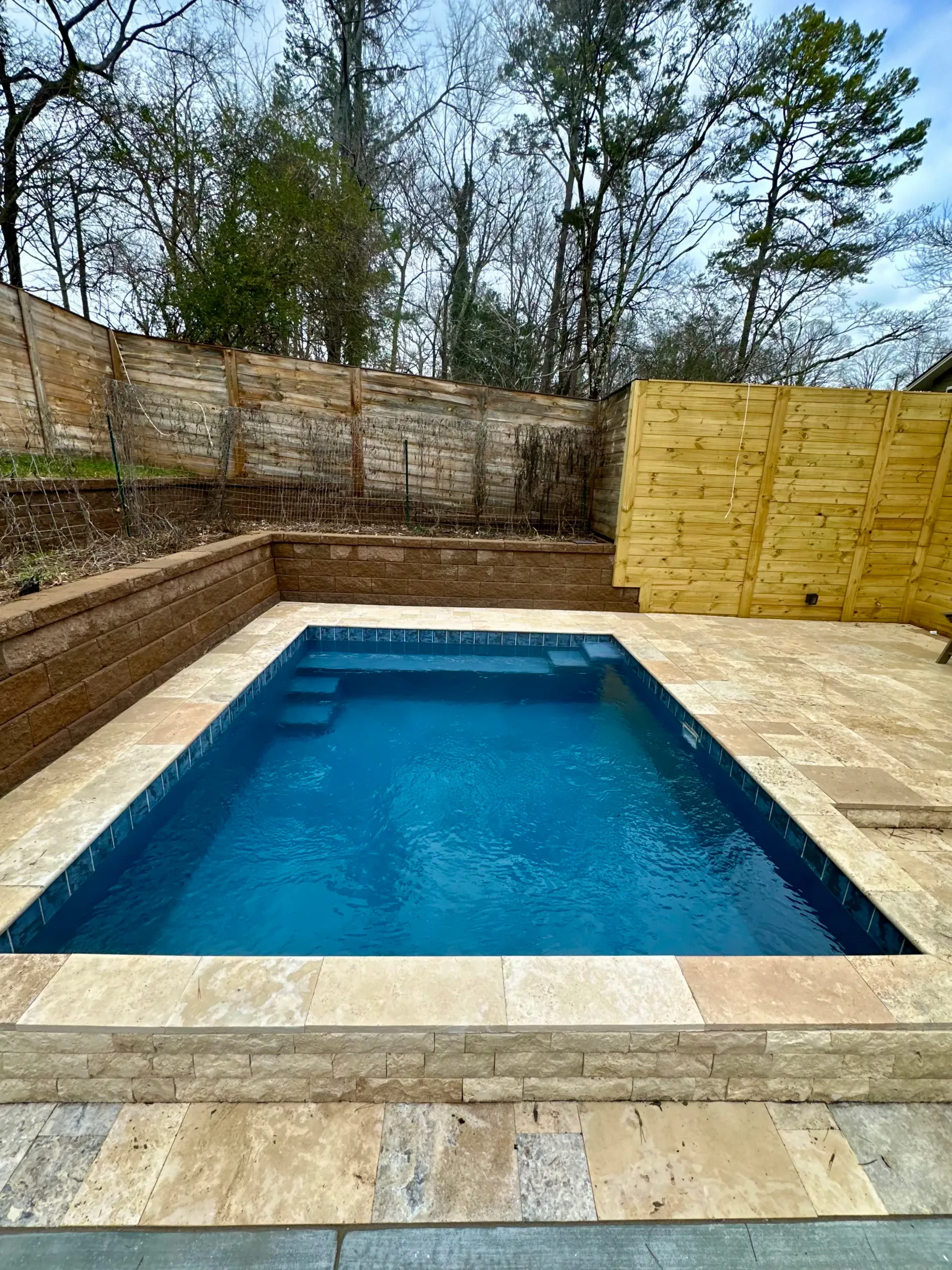 A small swimming pool surrounded by a brick wall and trees in a backyard.