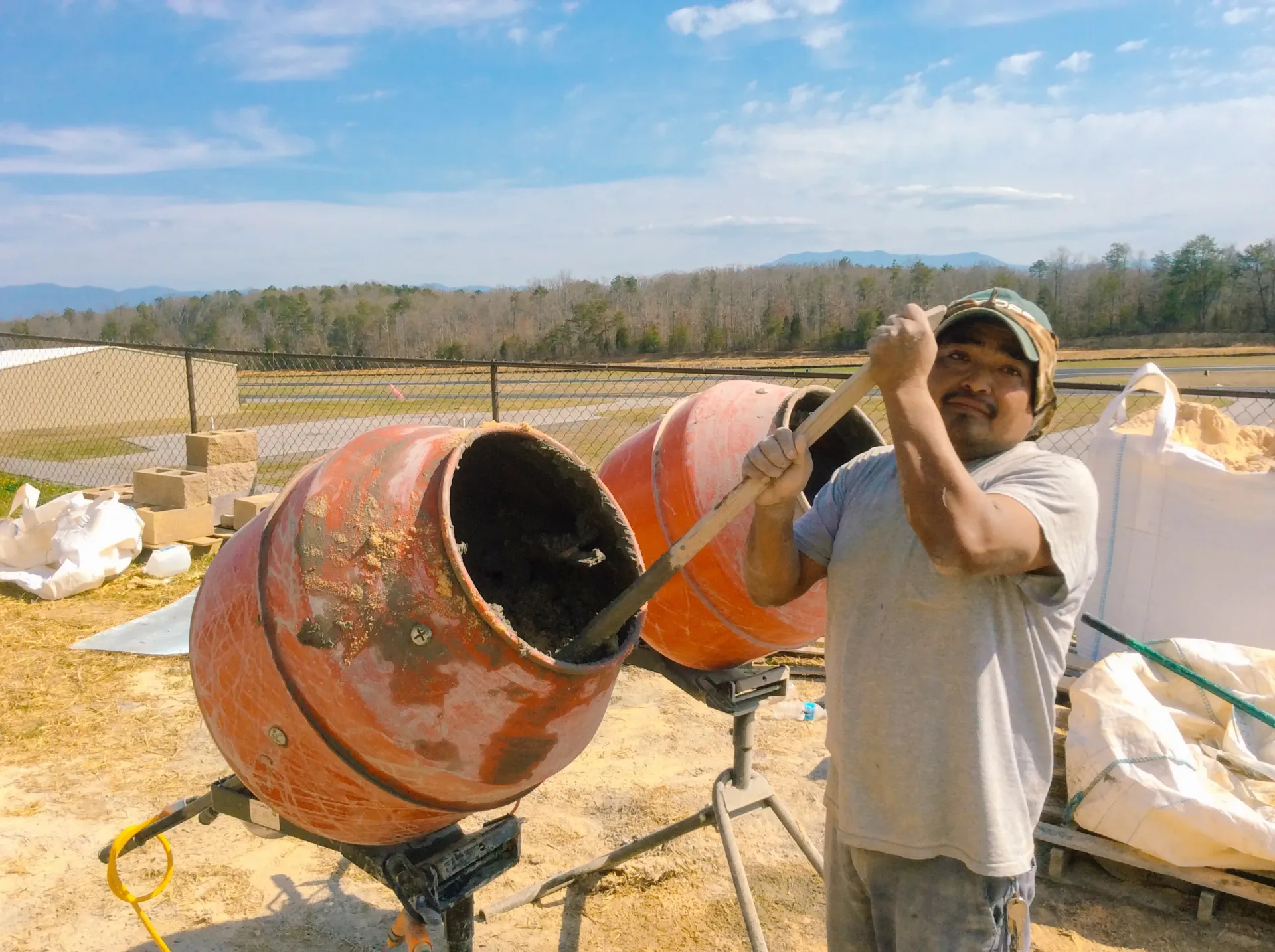 A man is standing in front of a cement mixer holding a shovel.
