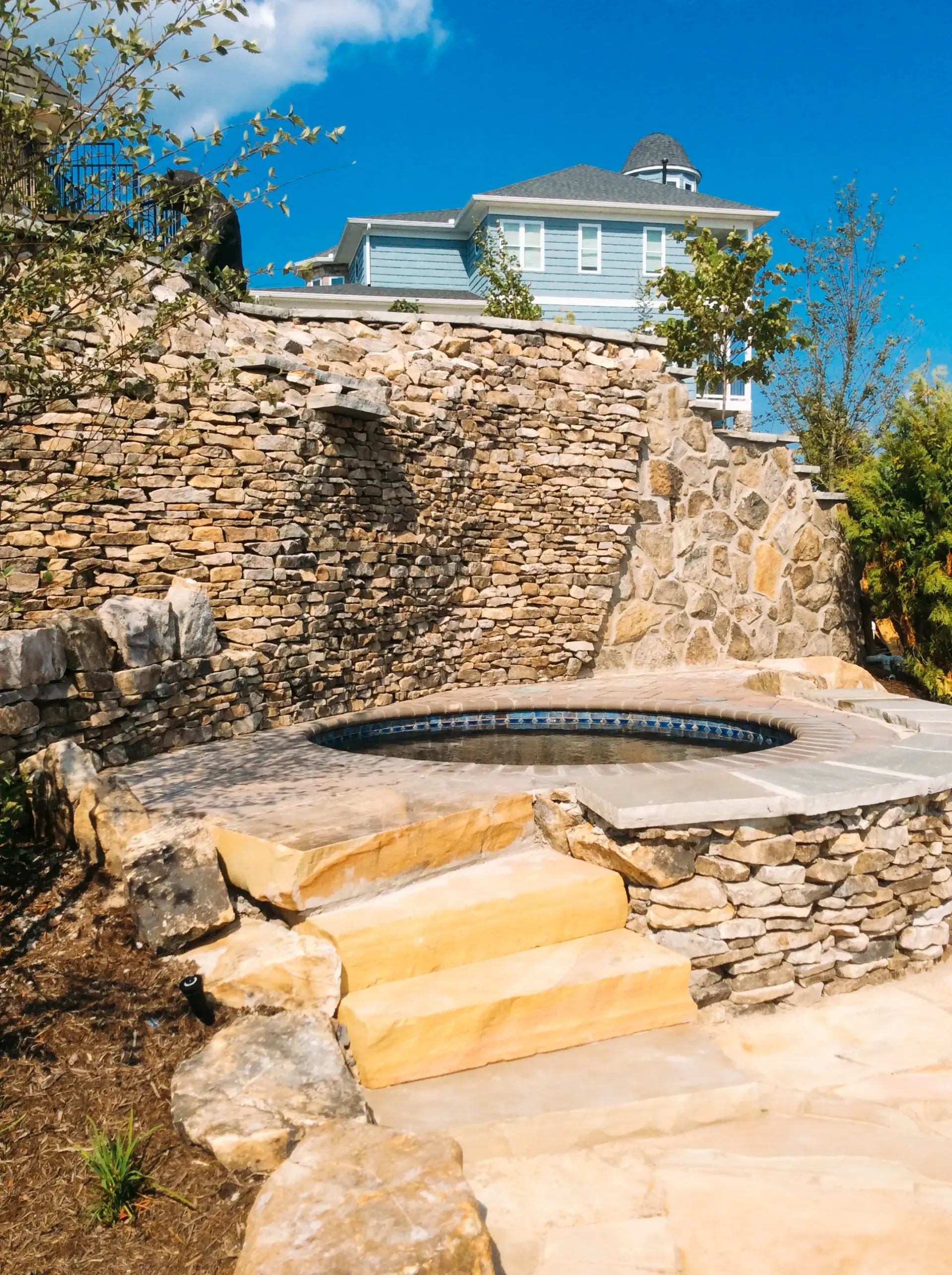 A stone wall with stairs leading to a hot tub in front of a house.