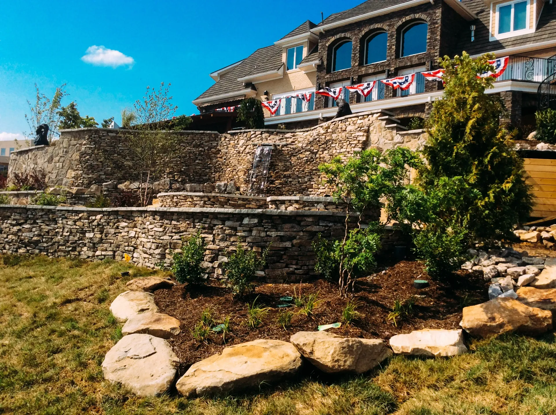 A large stone wall surrounds a lush green lawn in front of a large house.