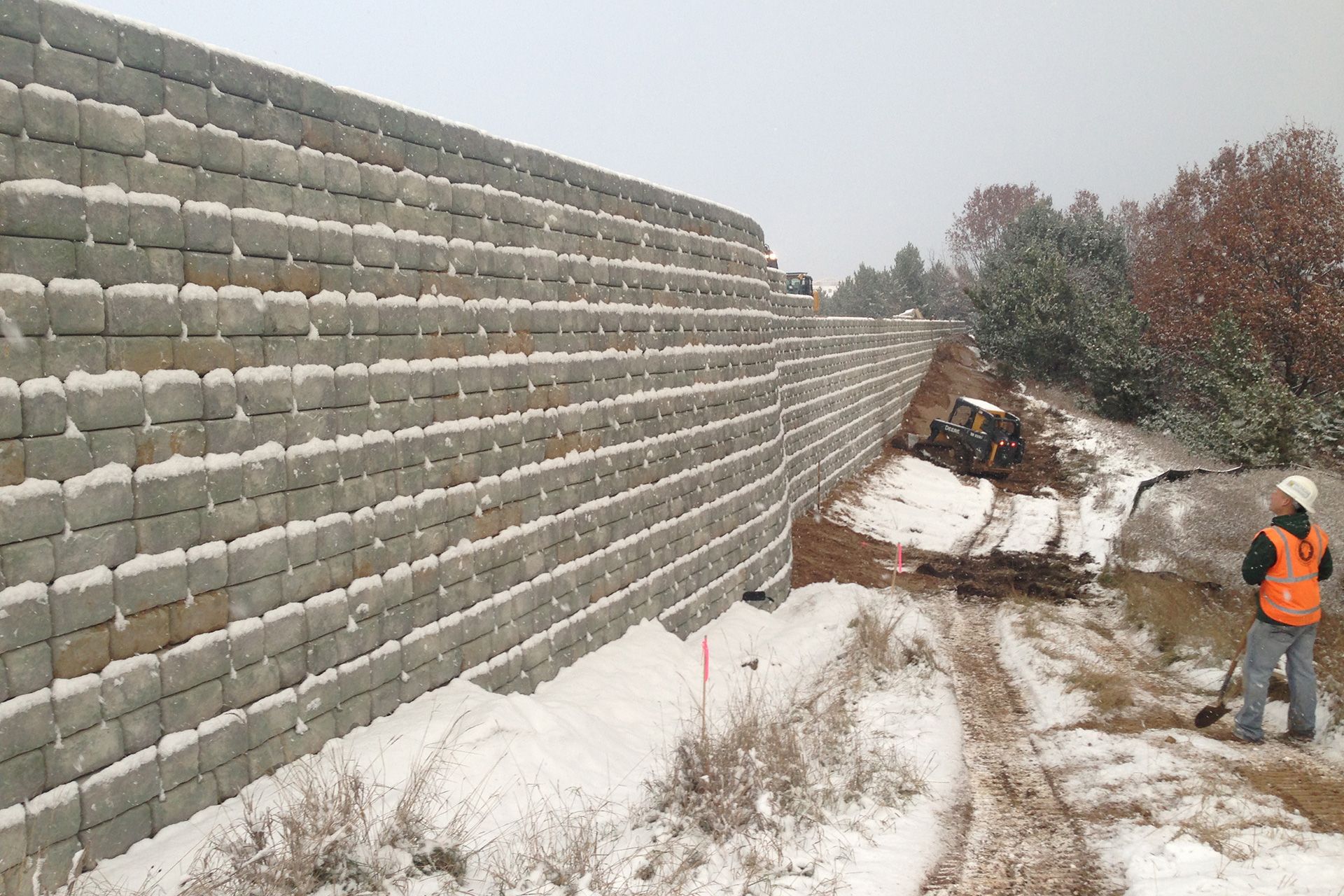A man in an orange vest is standing in front of a snow covered wall.