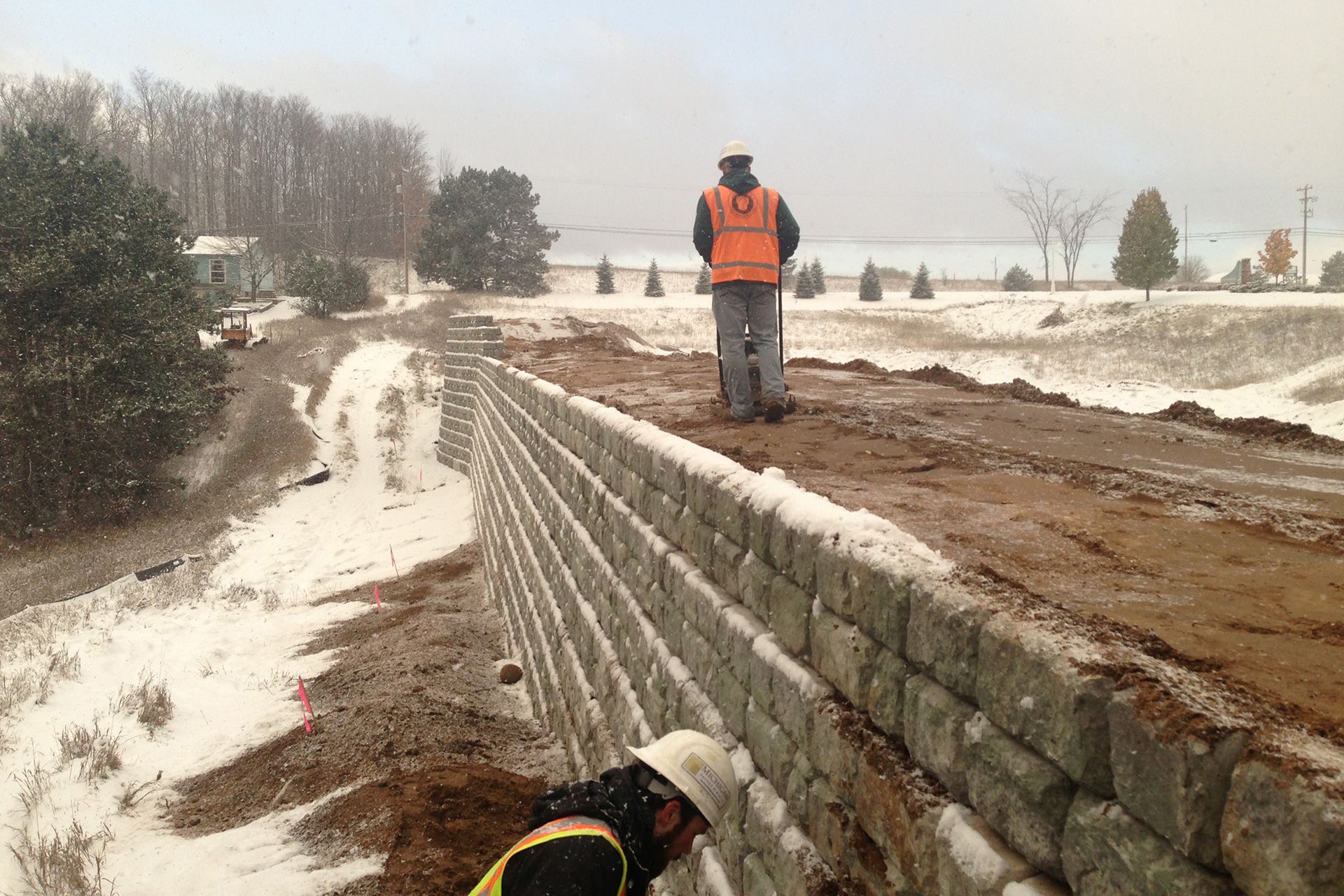 A man in an orange vest is standing next to a stone wall in the snow.