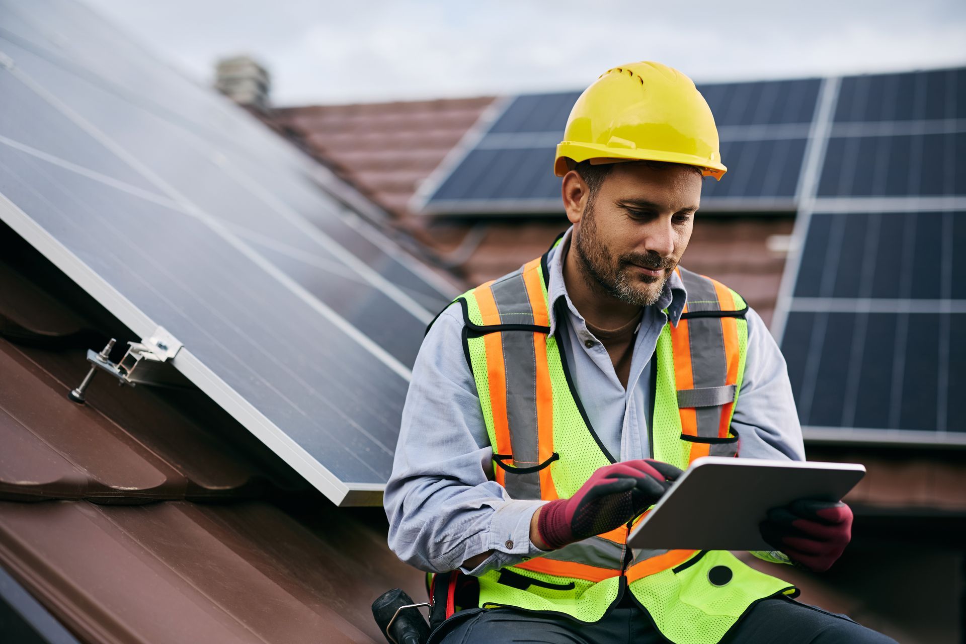 Solar panel technician on a roof with a tablet, wearing a safety vest and hard hat.