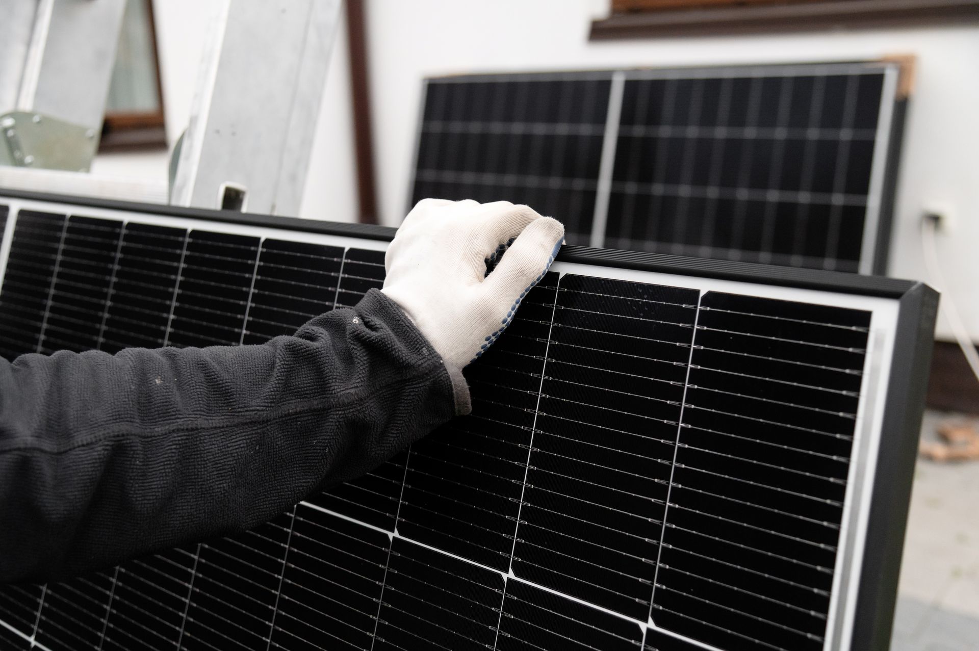 Gloved hand holding a black solar panel. Two more panels are in the background, indoors.