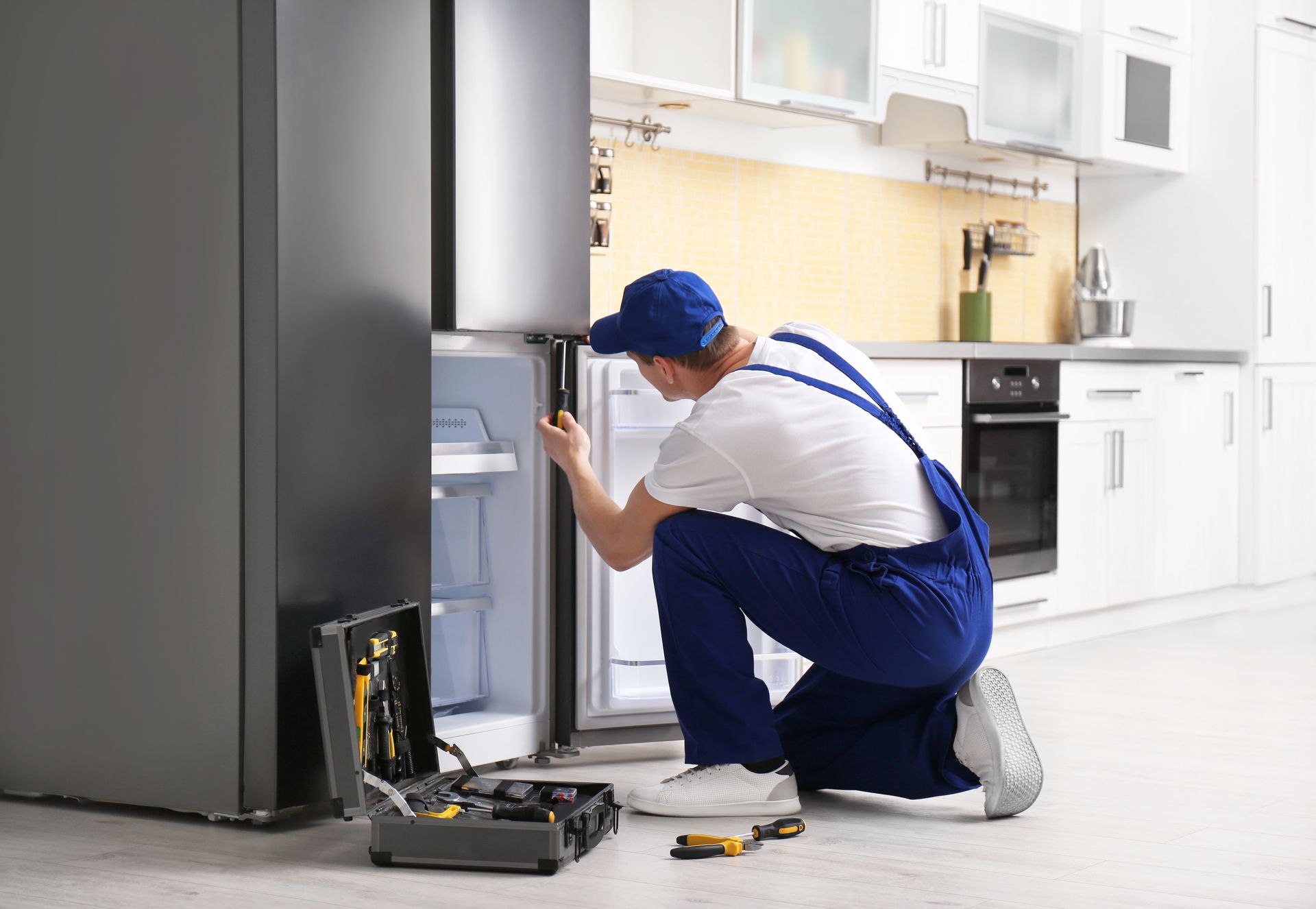 A refrigerator repairman in a blue uniform kneels, working on a fridge in a modern kitchen.
