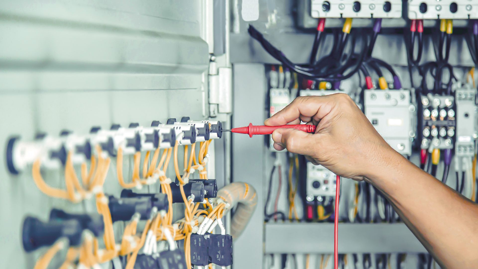 Hand testing electrical wiring in a gray control panel with a red multimeter.