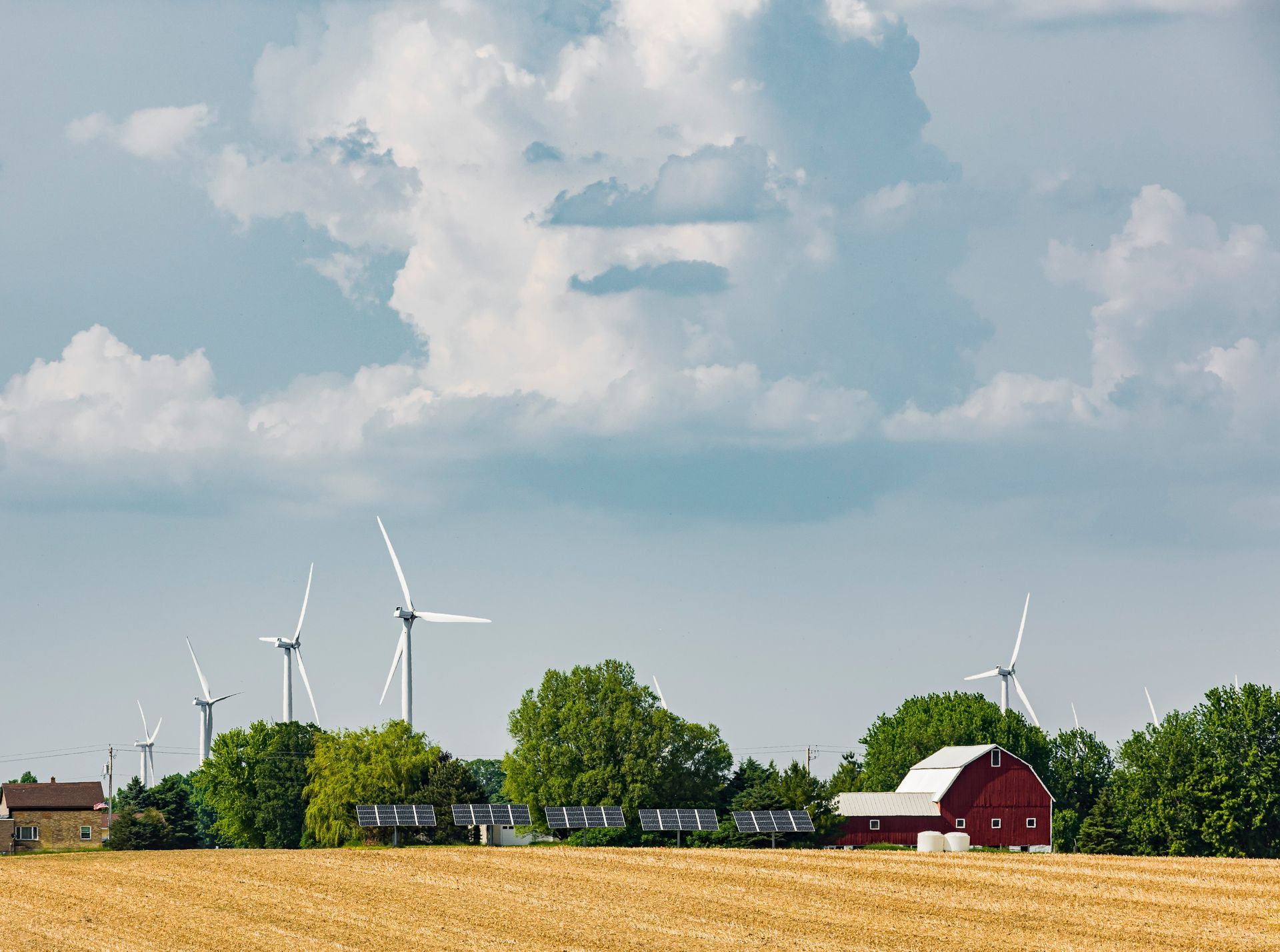 Wind turbines and a row of solar panels stand in a wheat field near a red barn under a cloudy sky.