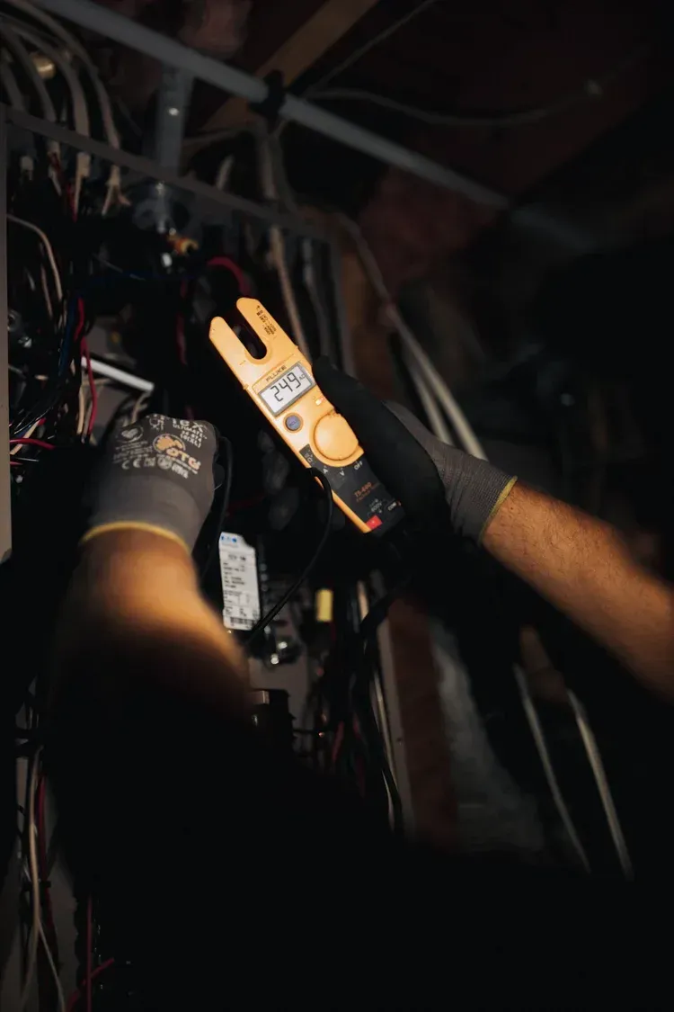 Electrician using a clamp meter to measure current in an electrical panel. Dark setting, wearing gloves.