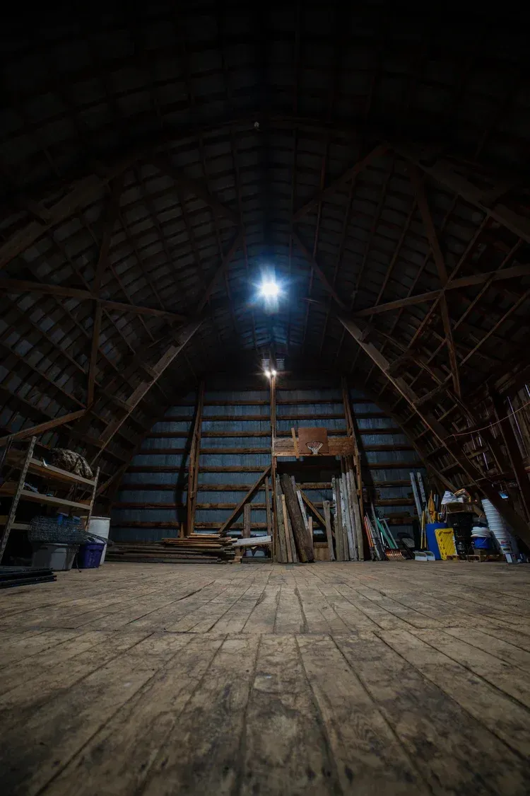 Interior of a dark barn attic, with a light source at the apex of the wooden structure and a wooden floor.