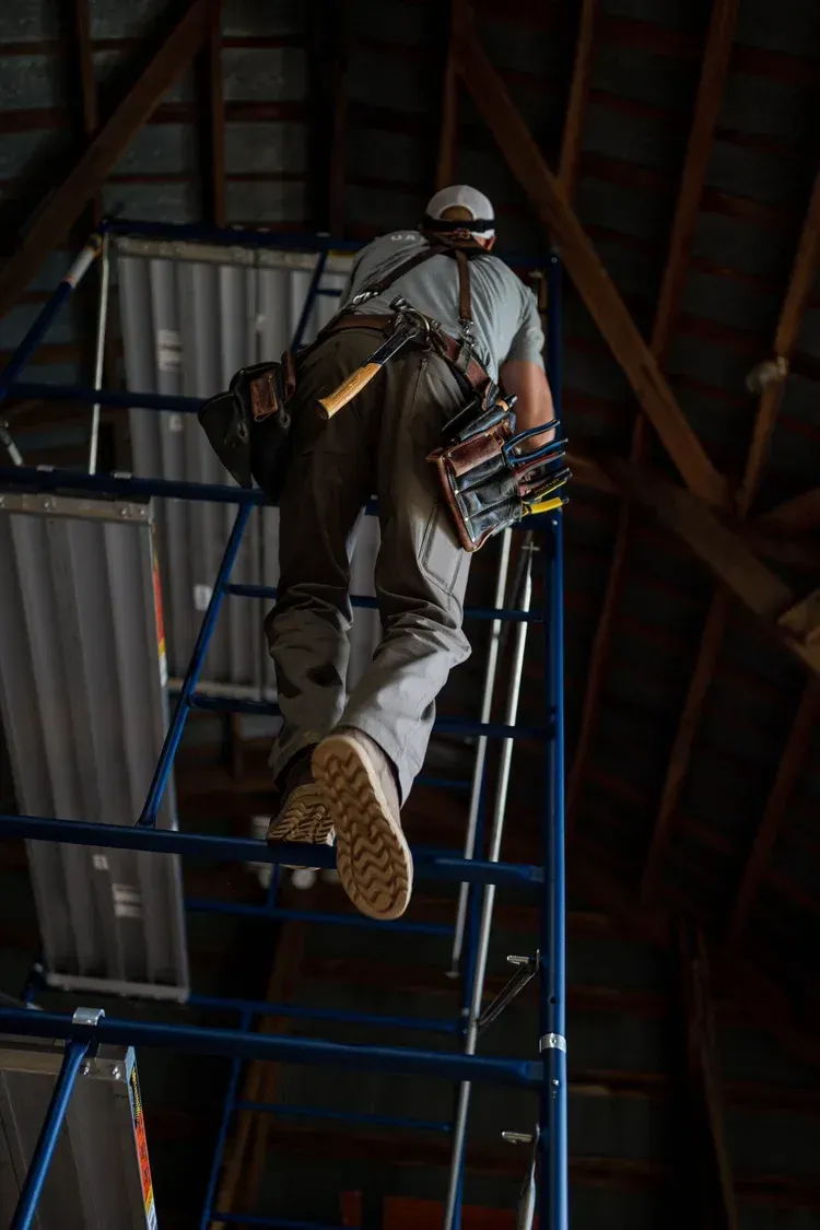 Person in work clothes climbing a scaffolding ladder inside a wooden building.