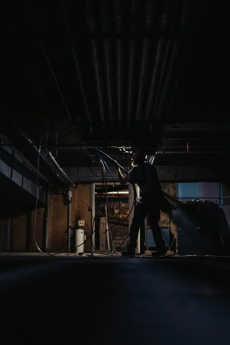 Person in a dark, industrial space, holding an object, possibly a weapon. Pipes and beams overhead.