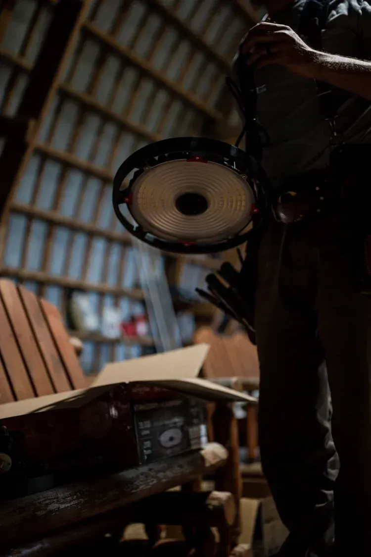 Man holding a banjo in a dimly lit, rustic interior with wooden furniture and a cardboard box.