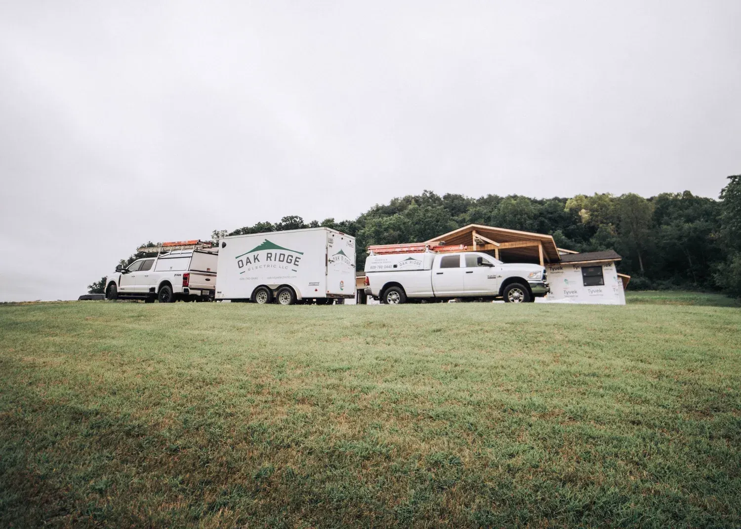Three white work trucks with trailer on a grassy hill; a partially constructed roof is in background.