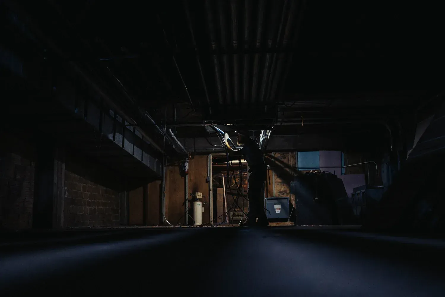 Dark, narrow basement. A person stands facing a doorway, under pipes and low ceiling.