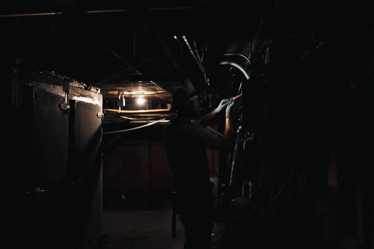 Man working in a dimly lit basement, reaching towards a fixture overhead.