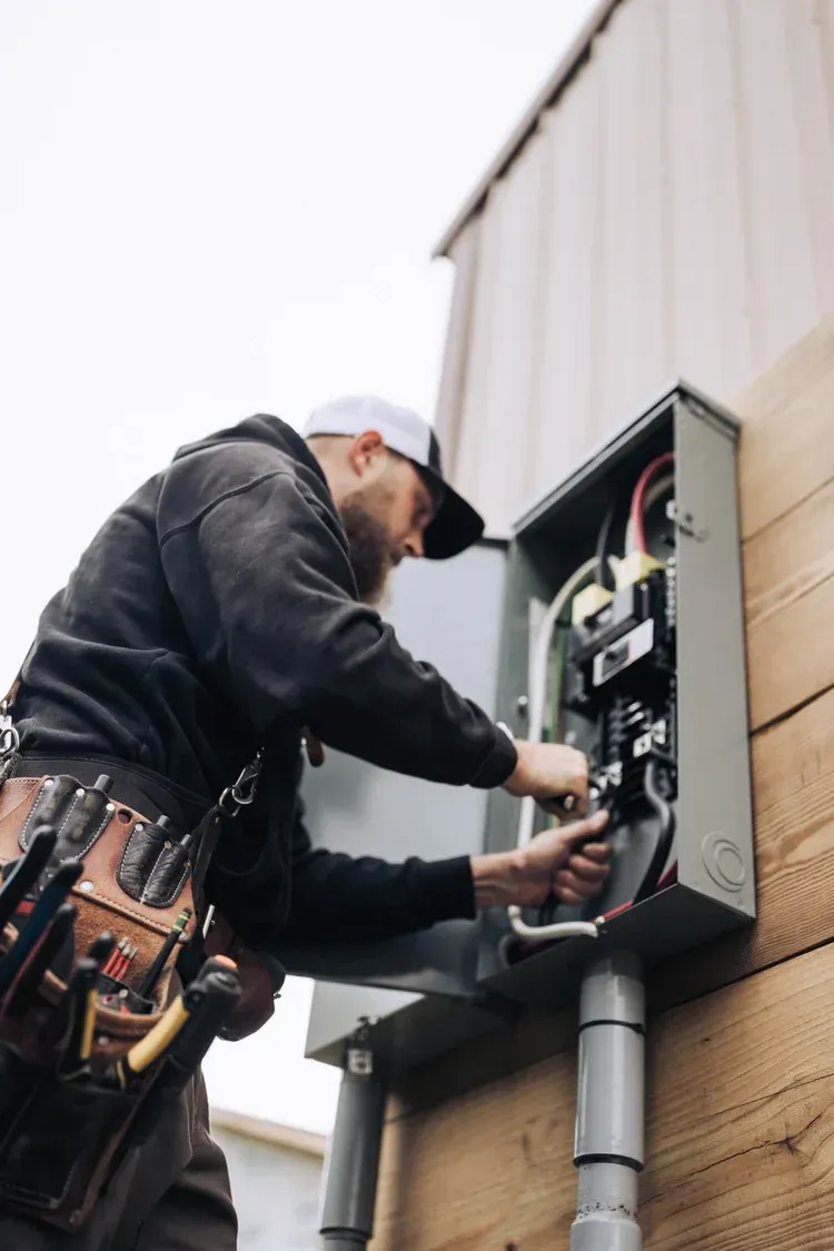 Electrician working on electrical panel, outdoors, wearing a tool belt and cap.