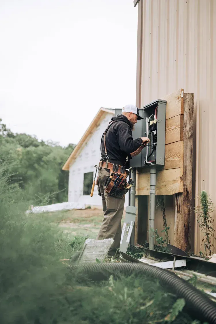 Electrician working on a gray electrical box mounted on the side of a building, a partially built house in the background.