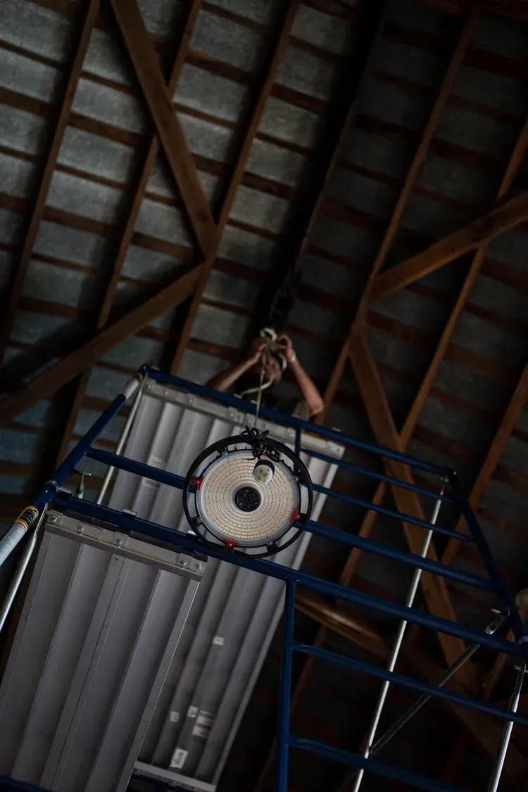 Person on a blue scaffolding adjusting a light fixture attached to a metal frame. Inside a wooden-beamed attic.