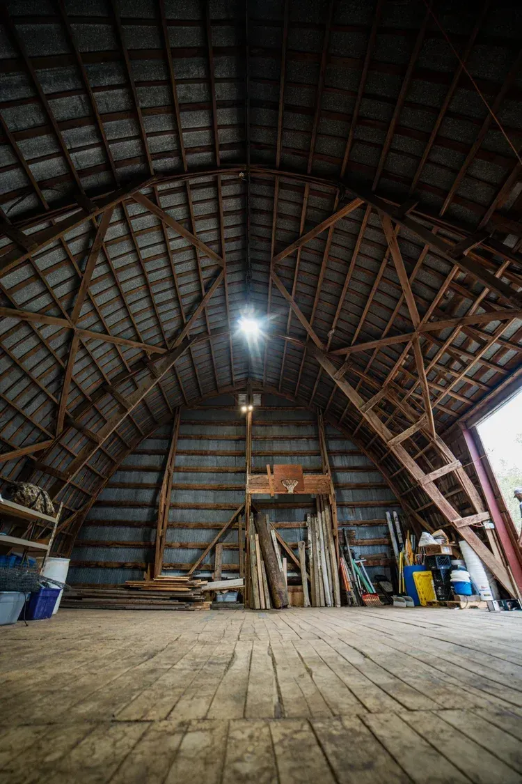 Interior of an old barn with wooden beams and a peaked roof. Sunlight shines down, illuminating the interior.