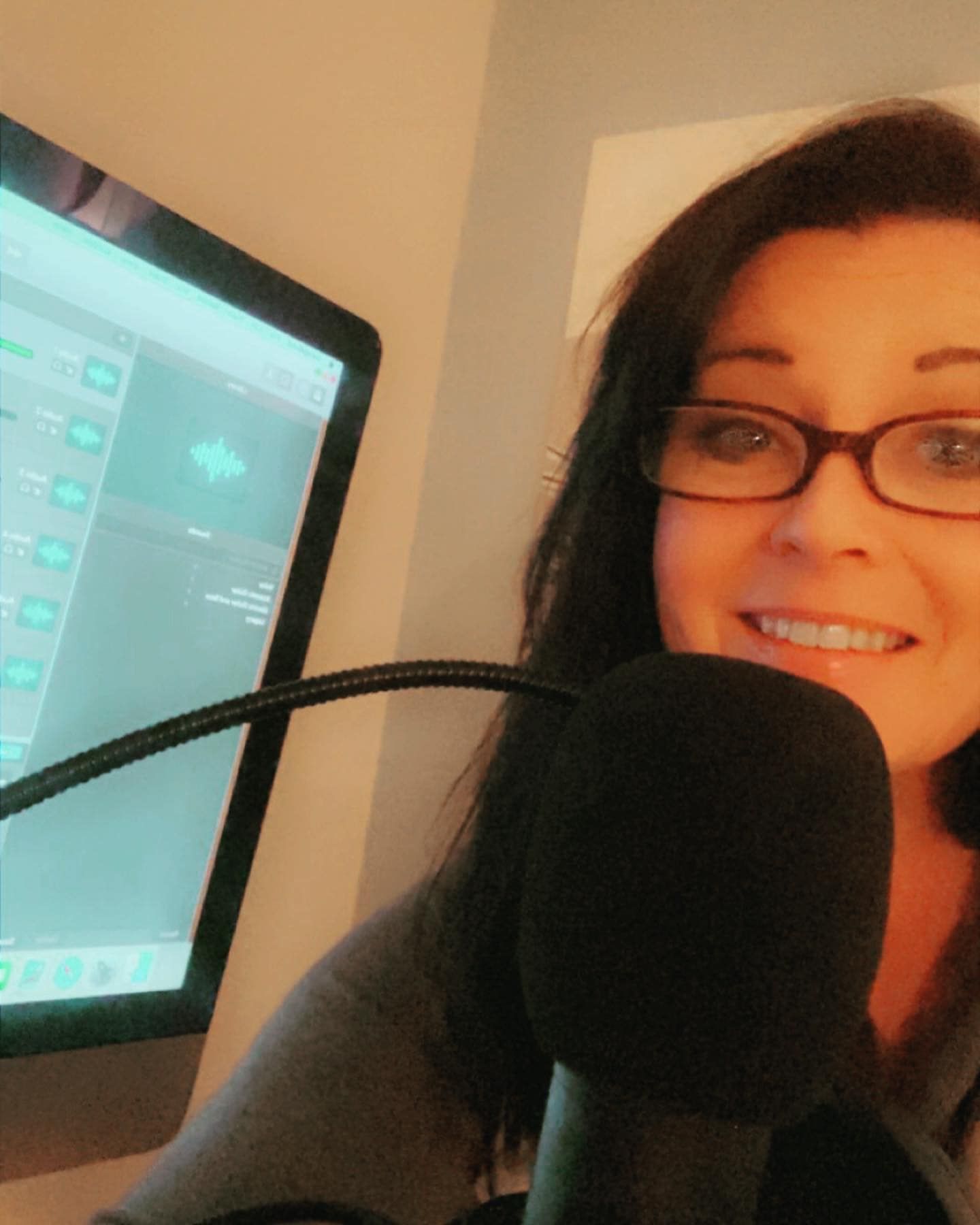Woman with glasses, smiling, recording a podcast with a microphone and computer.