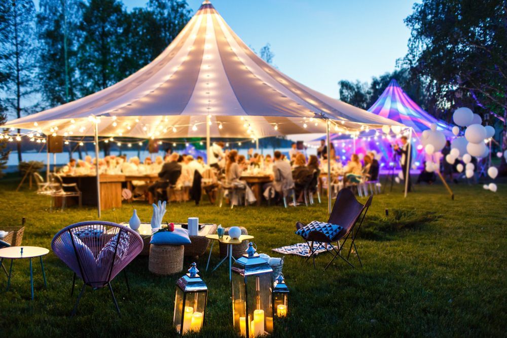 Outdoor evening tented party with white canopy, string lights, lanterns, and guests seated on the lawn