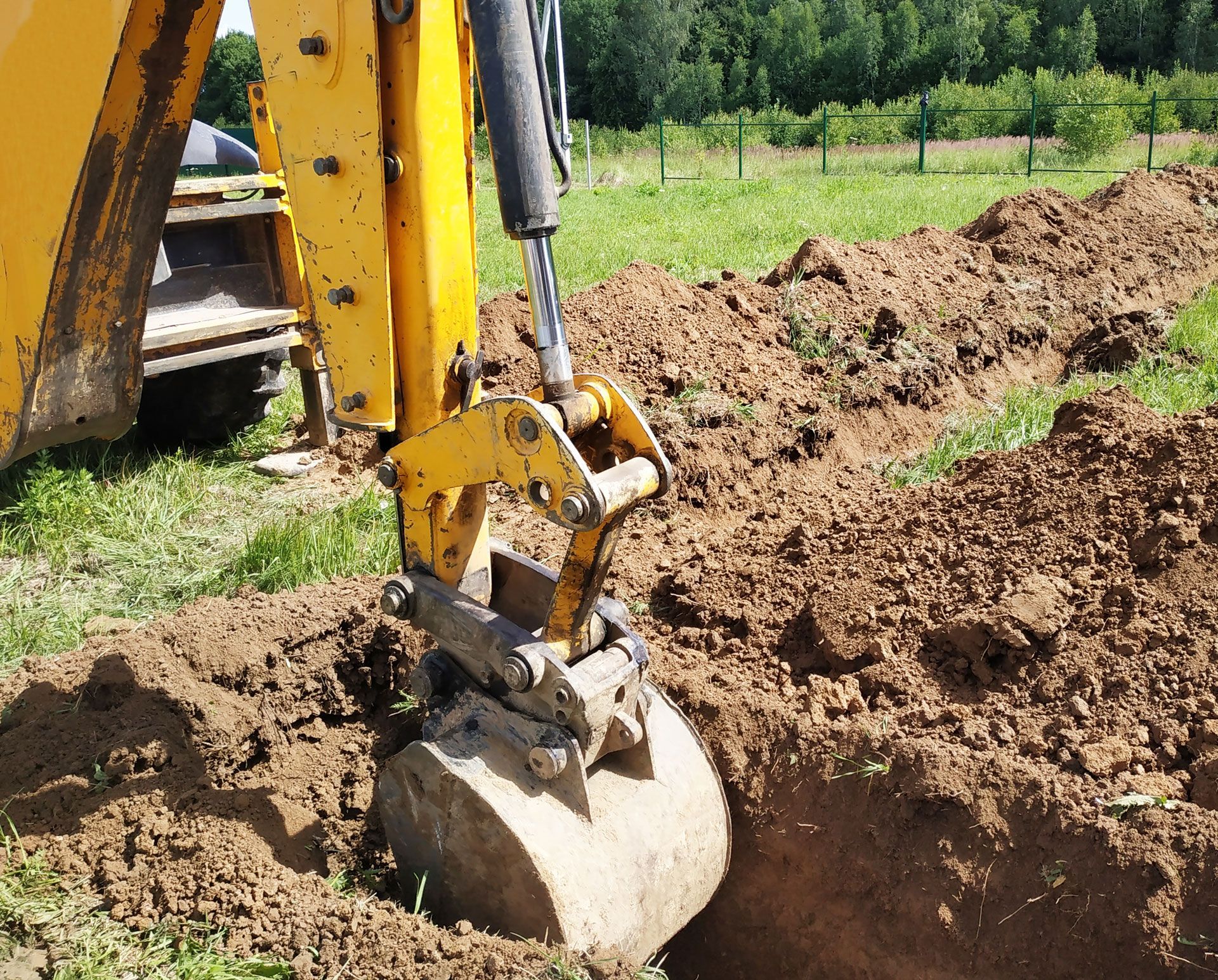 Excavator bucket digging soil in a dirt field beside green grass and trees