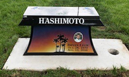 Gravestone with “HASHIMOTO” in white lettering and a sunset memorial plaque on a grassy lawn.