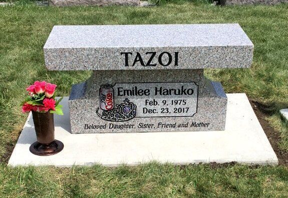 Gravestone with the name TAZOI and a memorial plaque, with flowers on the left.