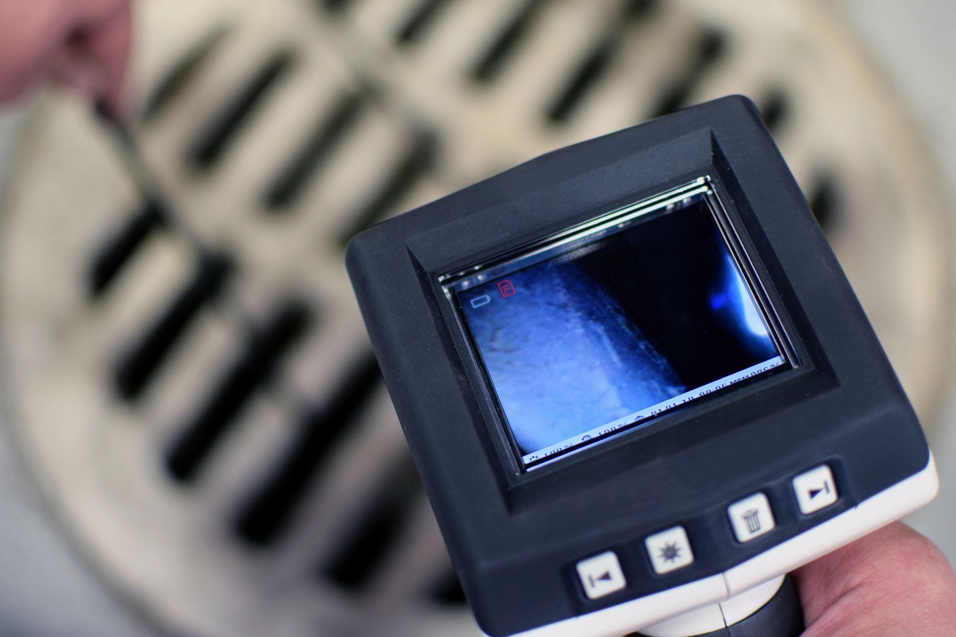 A handheld sewer camera monitor displays a dark pipe interior while positioned over a circular drain grate.